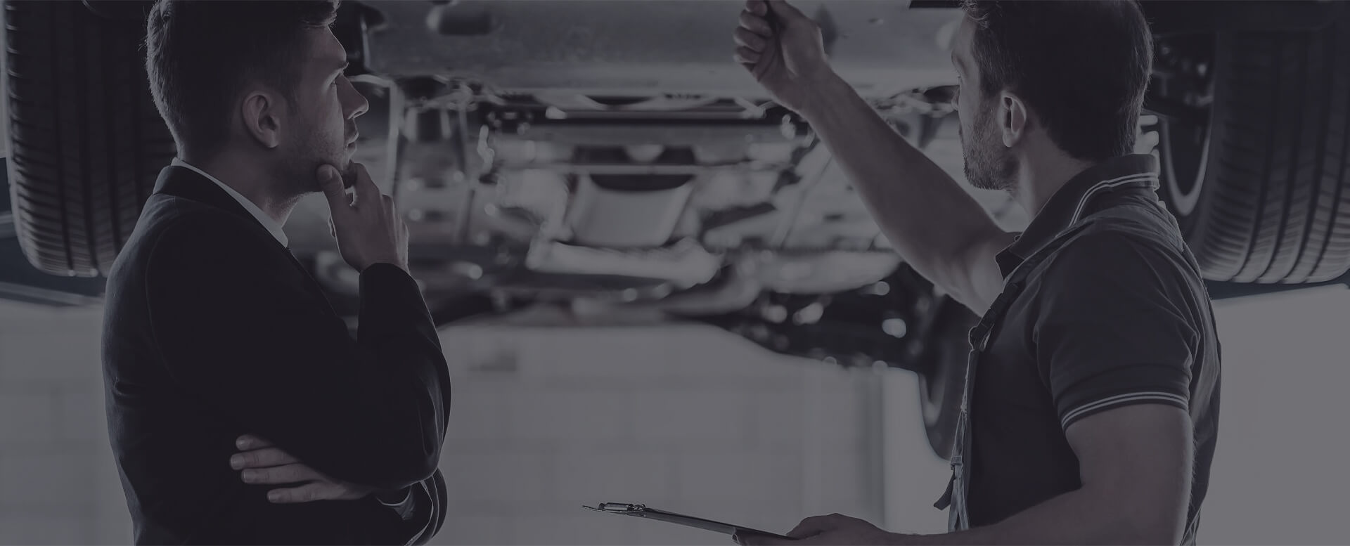 Zwei Männer, einer im Anzug und einer in Arbeitskleidung, stehen unter einem Auto und besprechen technische Details.